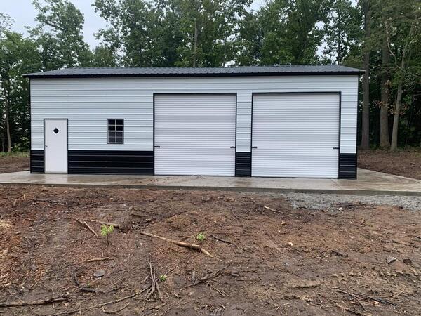 White metal garage with black lower panels, two roll-up doors, a walk-in door, and a window, on a concrete slab